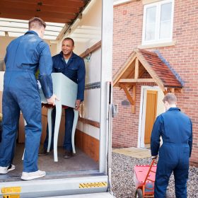 Removal Company Workers Unloading Furniture And Boxes From Truck Into New Home On Moving Day